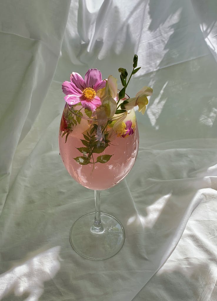 A stylish pink drink garnished with fresh flowers in a wine glass under natural light.