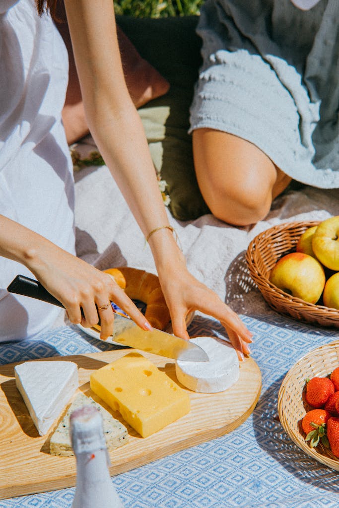 A sunny picnic scene with hands cutting cheese, surrounded by apples and strawberries.