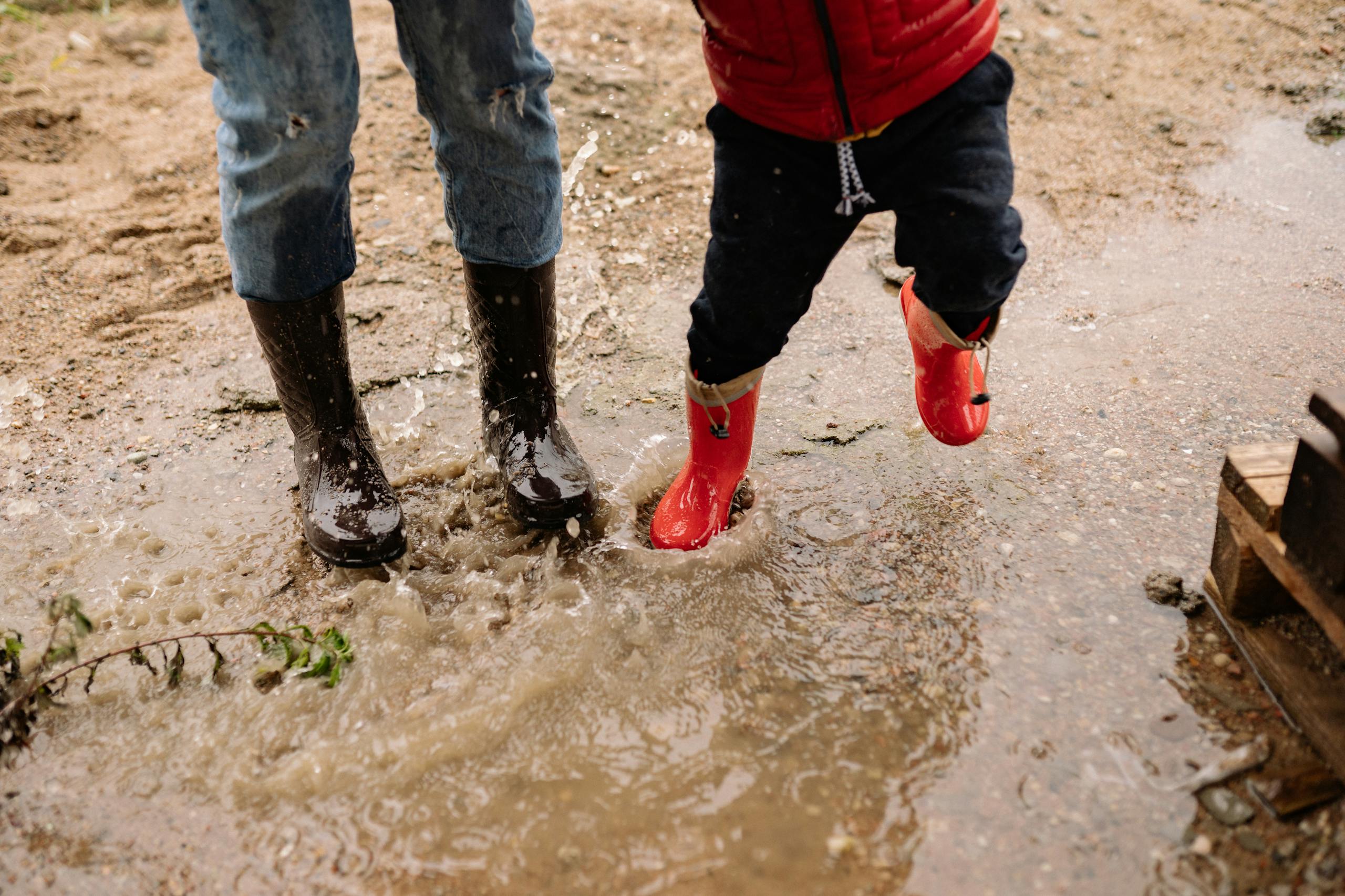 Two kids jumping and splashing in muddy puddles wearing black and red wellington boots.