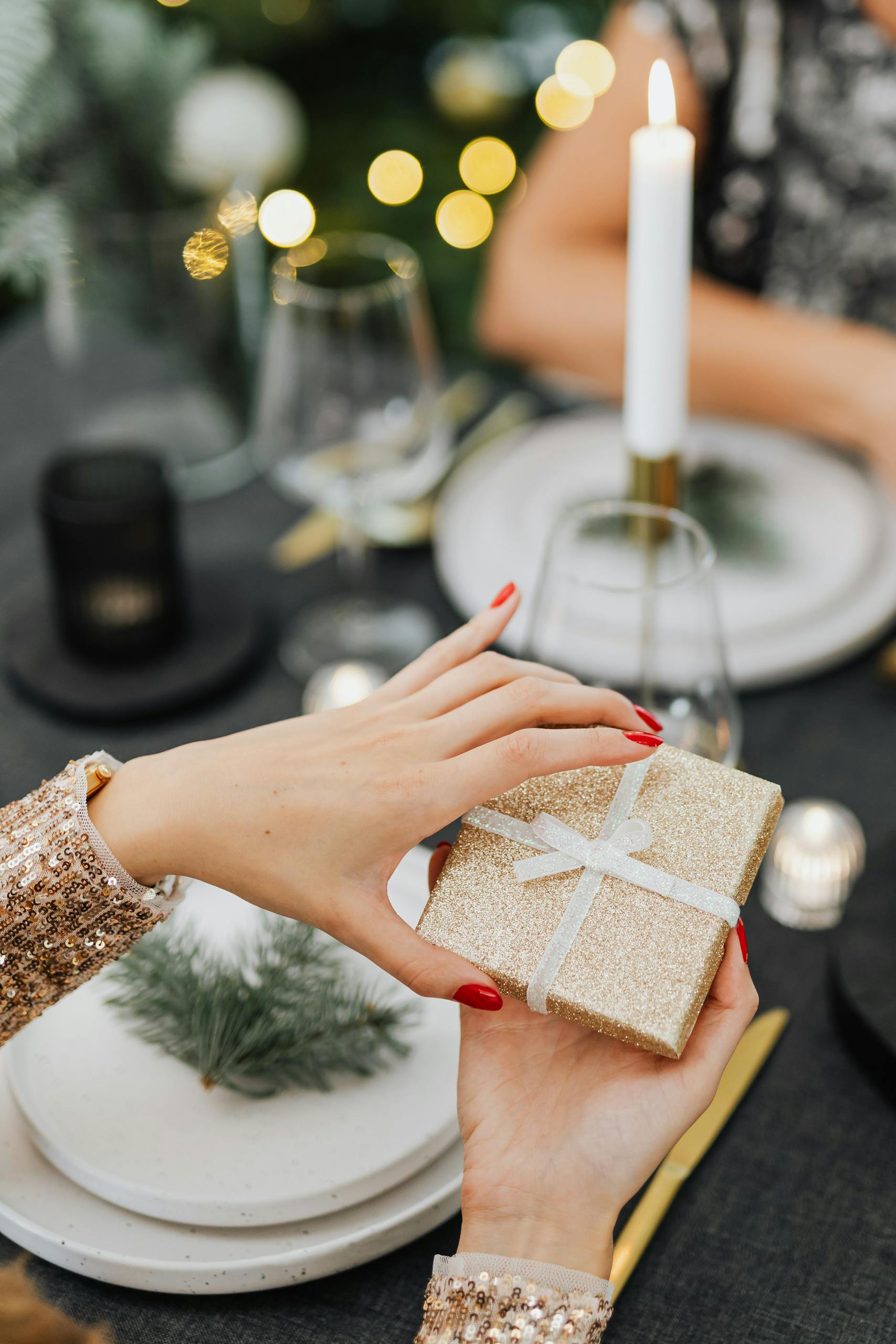 A close-up of two women exchanging a glittery Christmas gift at a candlelit dinner table.