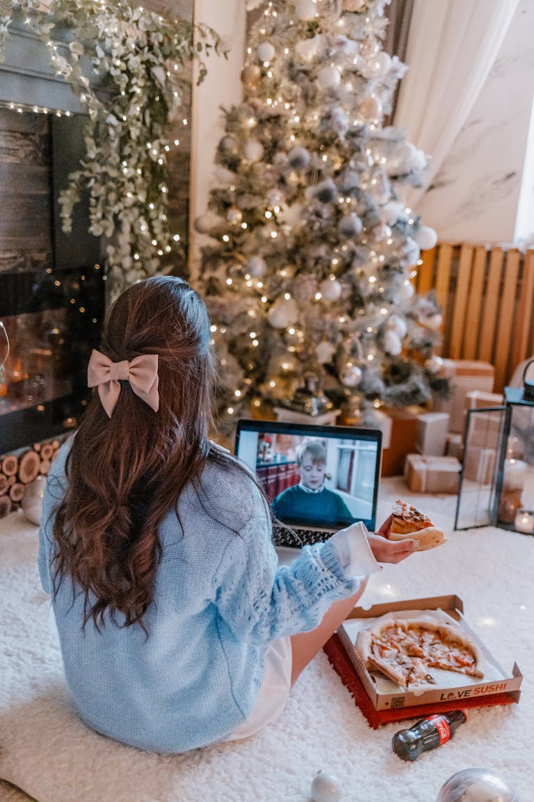 Cozy home alone Christmas vibes with pizza and laptop near decorated tree.