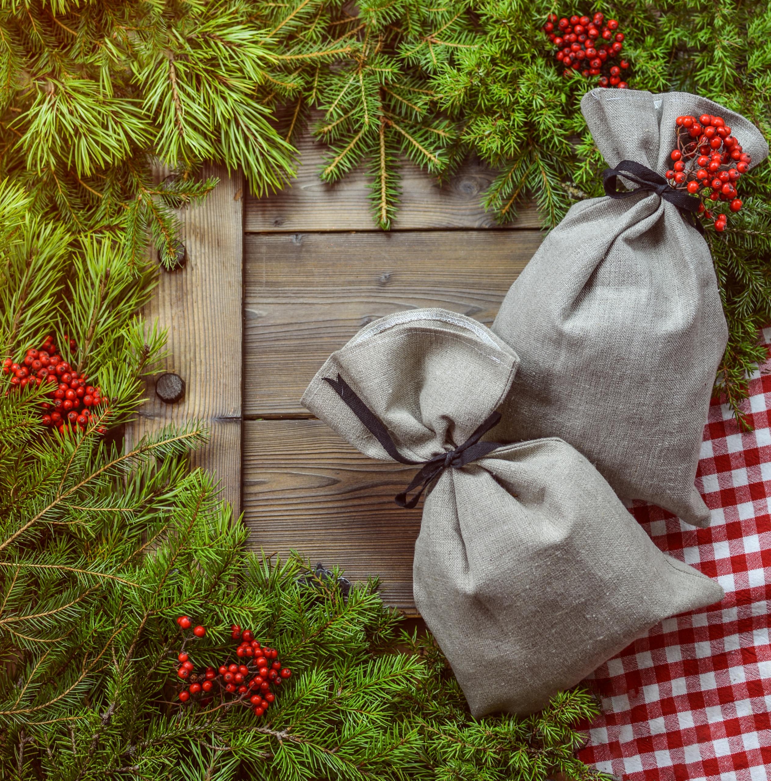 Festive linen gift bags on a wooden table surrounded by evergreen branches and red berries, perfect for holiday themes.