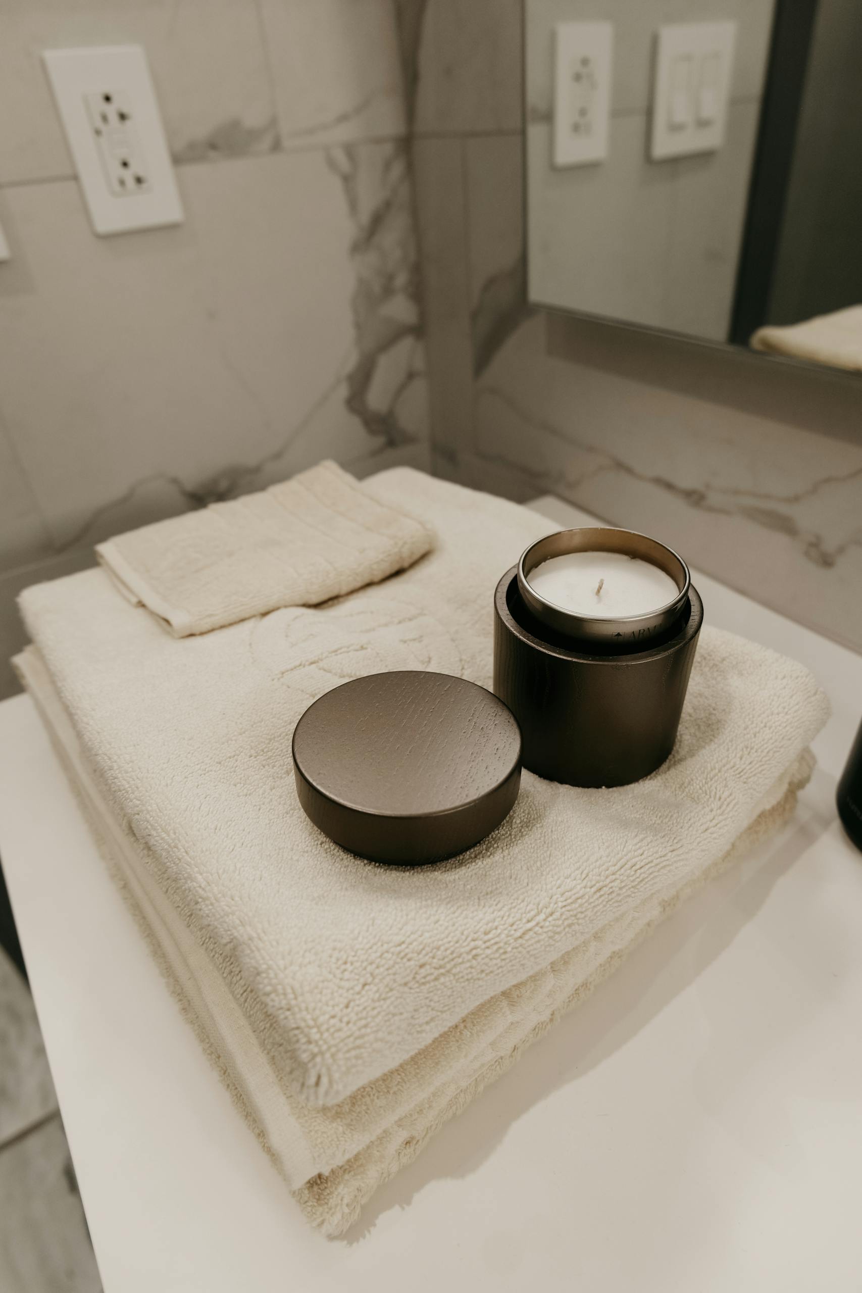 A modern bathroom setting featuring stacked white towels and a black candle on a counter.