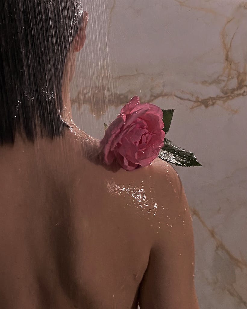 Back view of a woman showering with a pink rose on her shoulder, water cascading down. perfect for an autumn self care Sunday