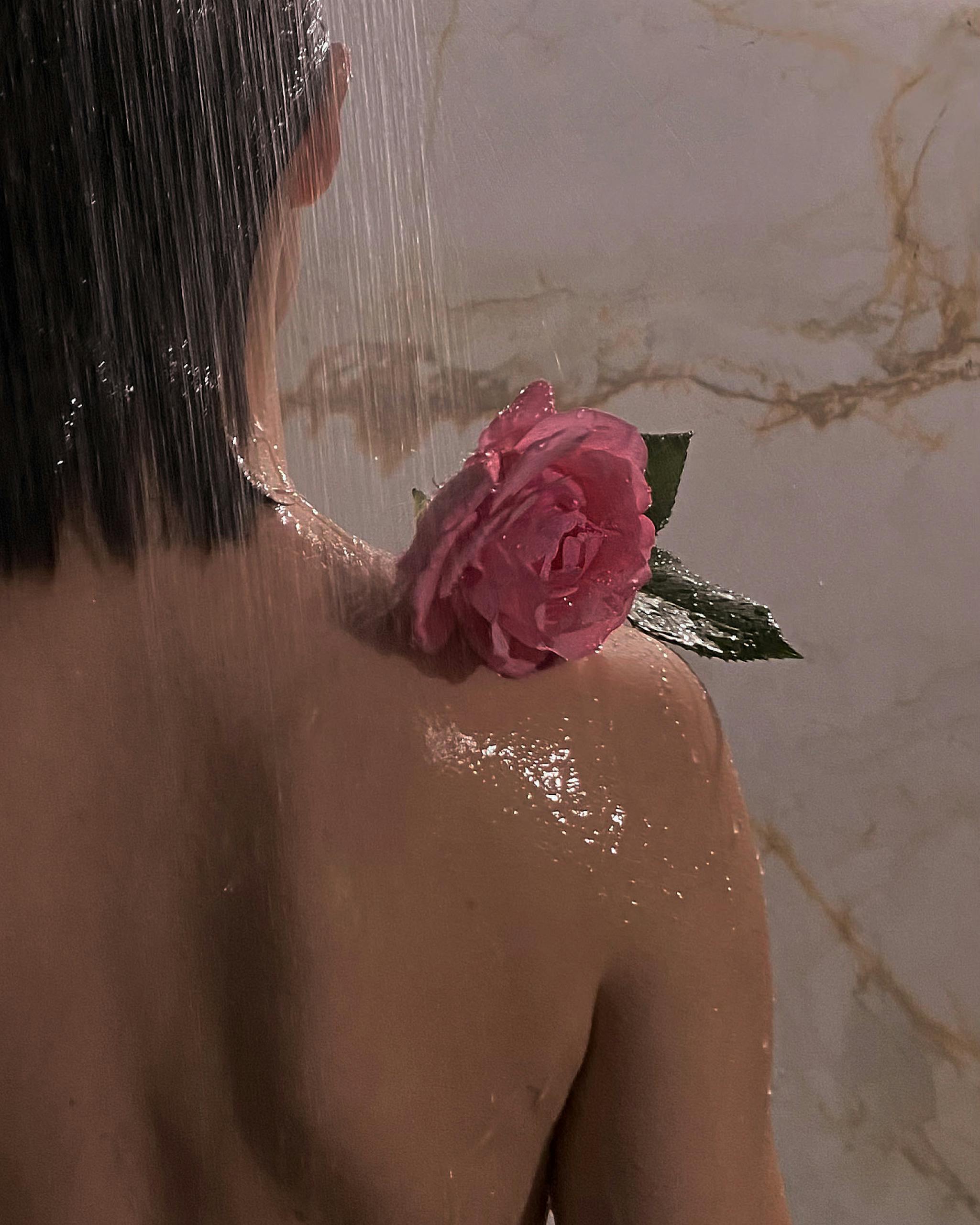 Back view of a woman showering with a pink rose on her shoulder, water cascading down.