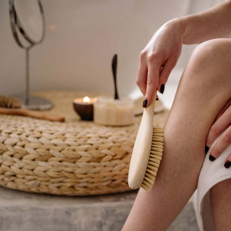 Close-up of a woman using a dry brush for skin exfoliation in a cozy bathroom setting.