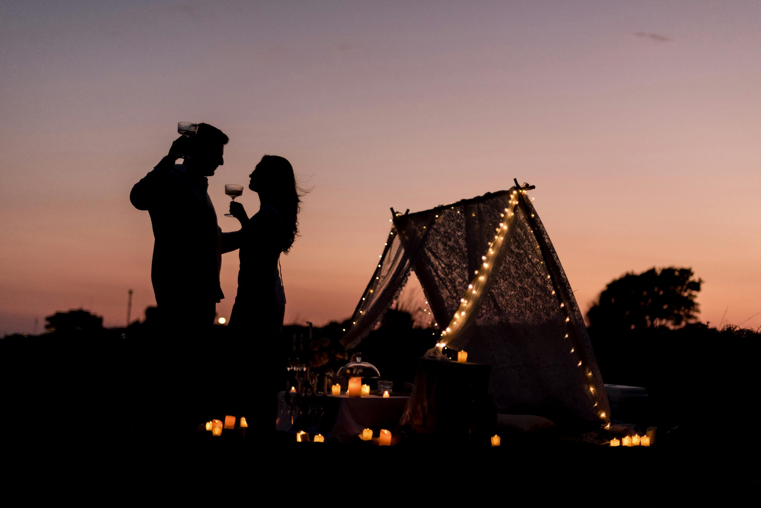 Silhouette of a couple toasting wine glasses during a romantic sunset camping sleepover setup with candles.