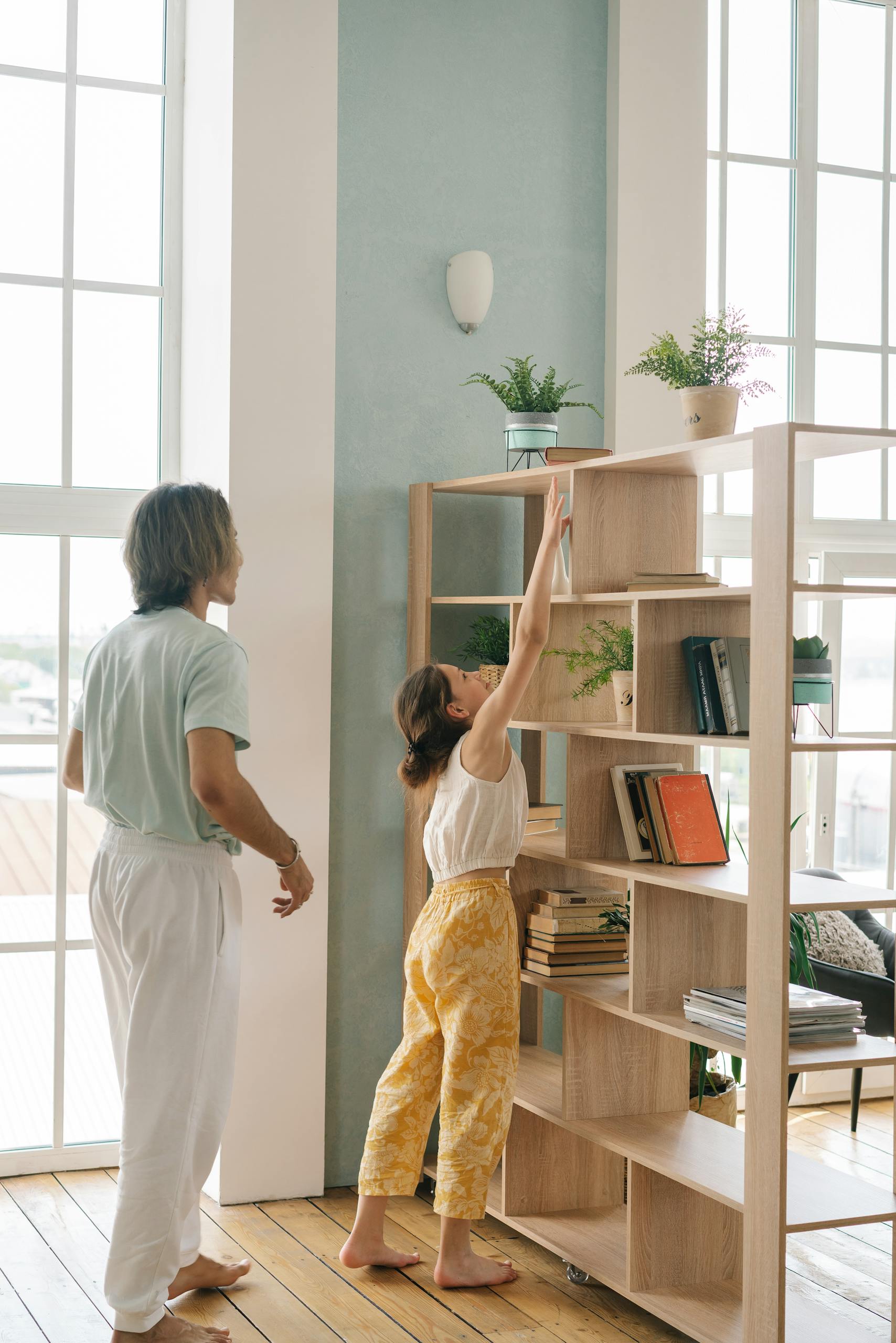 A father and daughter bonding while reaching for books on a shelf in a cozy, well-lit living room.