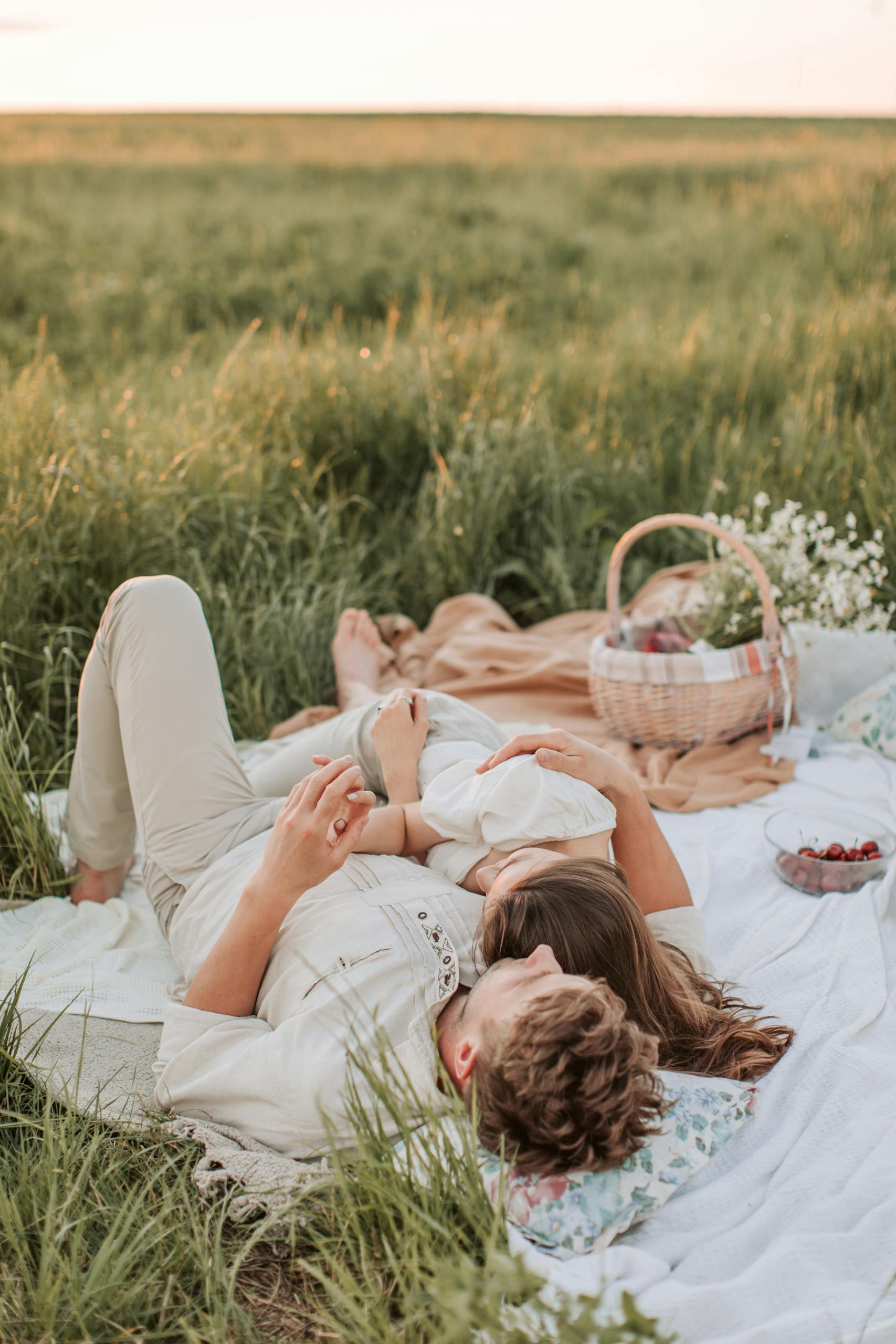 A couple enjoys a romantic picnic outdoors, lying on a blanket in a sunny grass field.