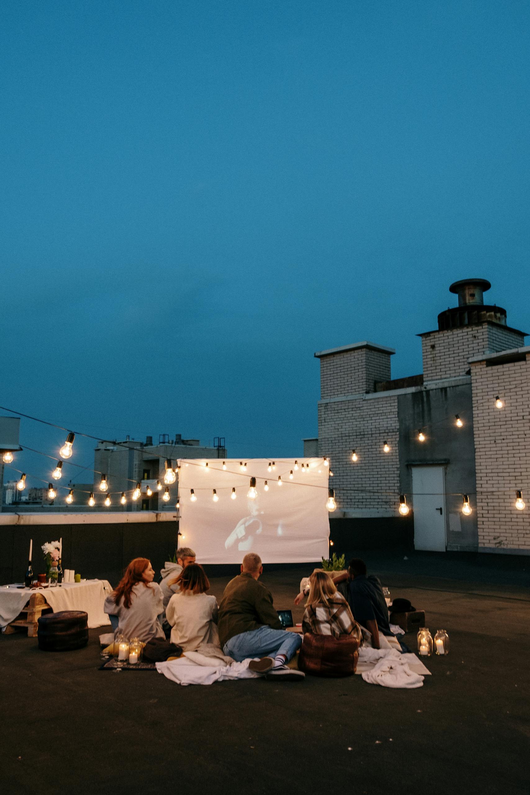A group of friends enjoys a rooftop movie night under string lights with a projector and cozy blankets.
