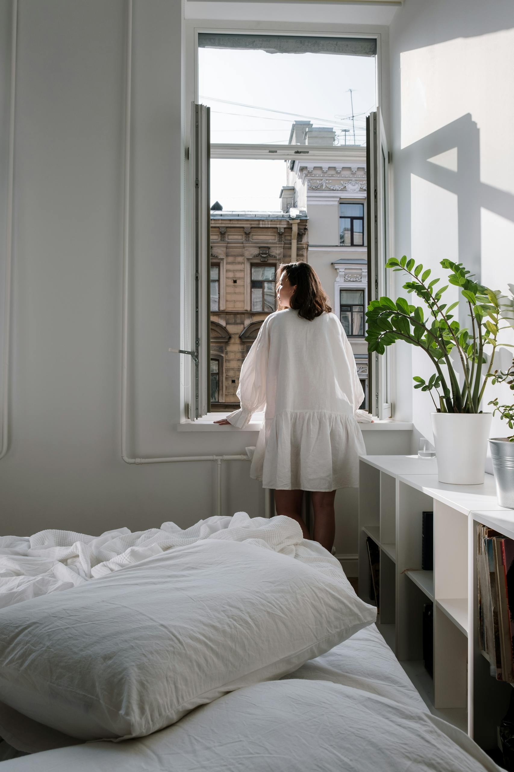 A woman gazes out the window of a bright, minimalist urban bedroom, surrounded by plants and soft bedding.