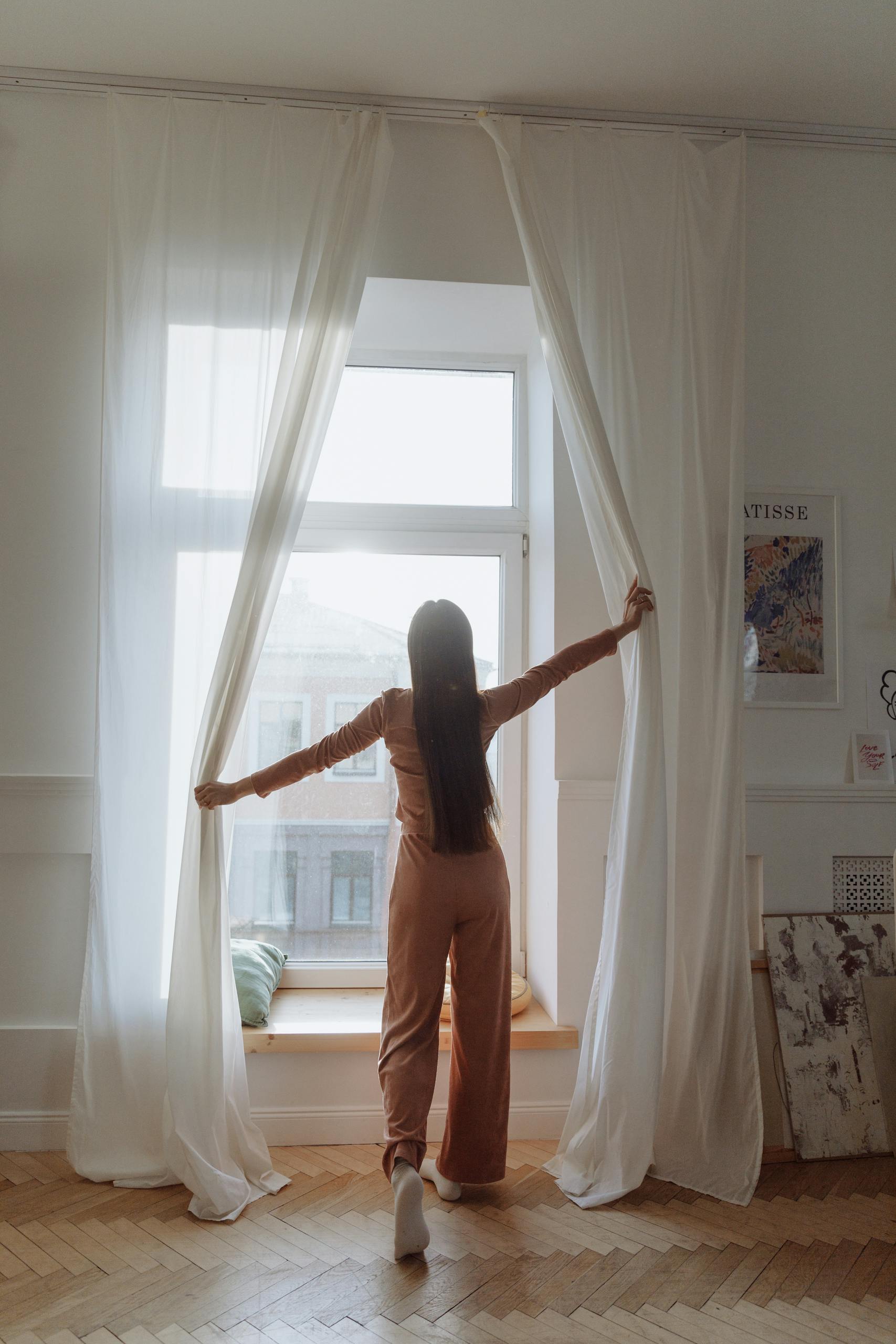 A woman stands indoors, opening curtains to let in the morning sun. Minimalist interior.