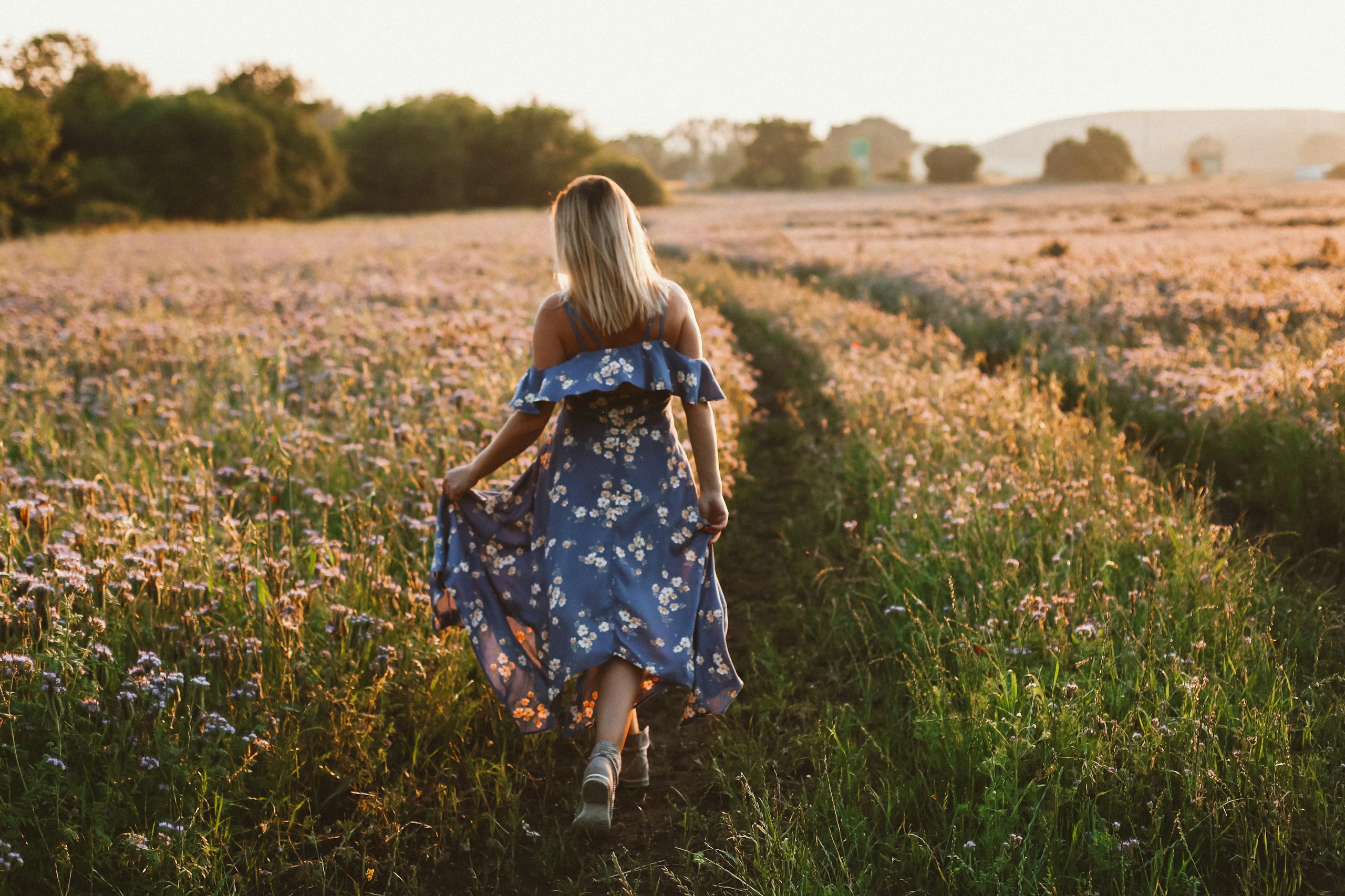 A woman walking in a floral dress through a bright flower field during sunset, showcasing tranquility and nature.