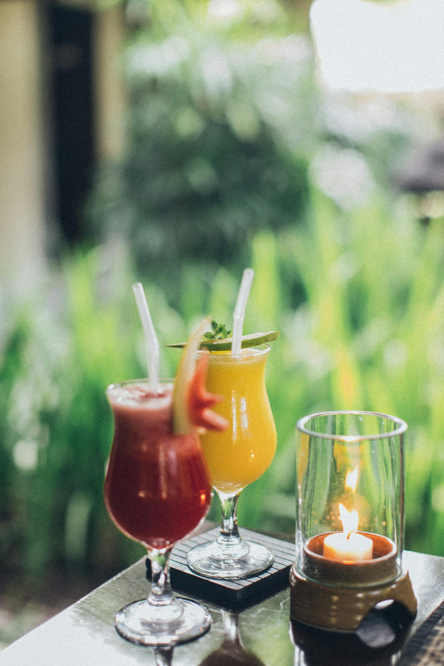 Vibrant cocktails adorned with straws on a garden table beside a glowing candle.