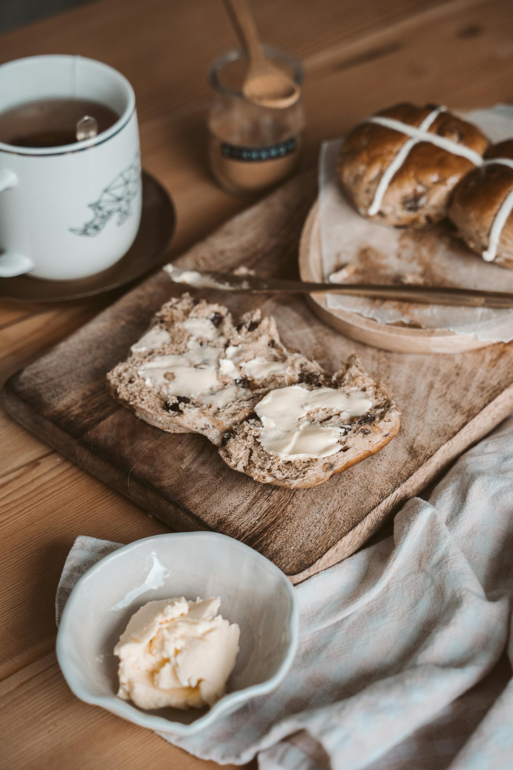 Warm rustic breakfast spread featuring hot cross buns, butter, and a cozy tea setup.