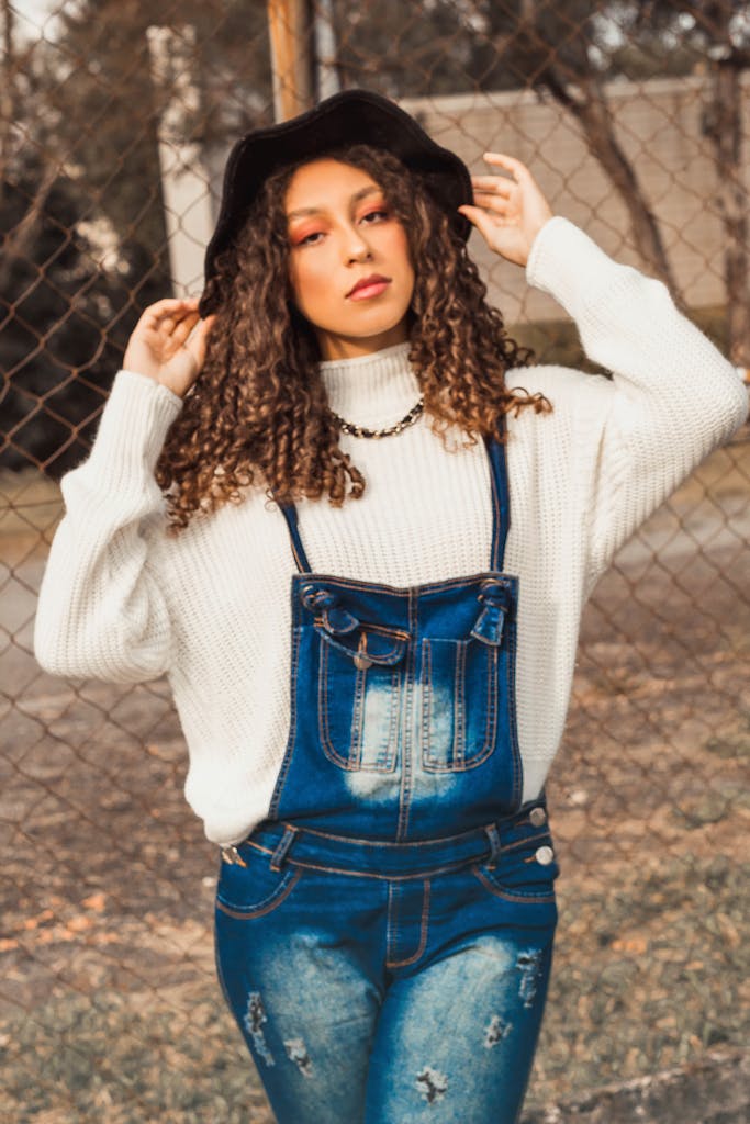 Fashionable woman in denim overalls and white sweater posing outdoors
