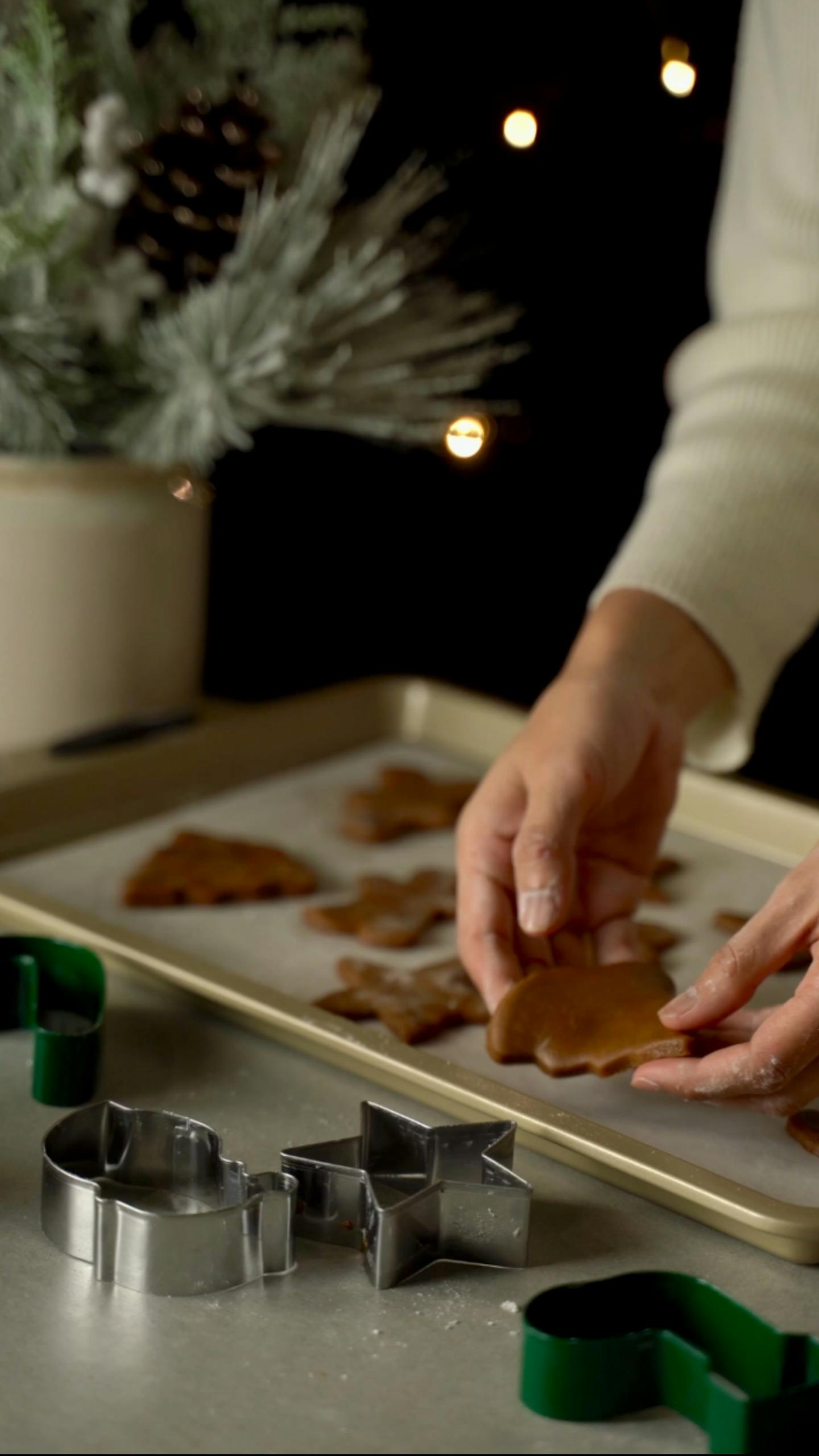 Hands preparing gingerbread cookies using cutters, set against a festive background with holiday decor.