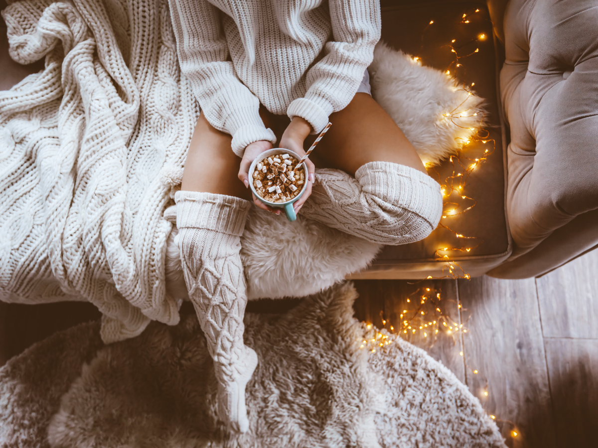 woman relaxing on sofa with warm drink surrounded by candles and fairy lights in softly lit living room — how to make your home feel like a permanent escape every day
