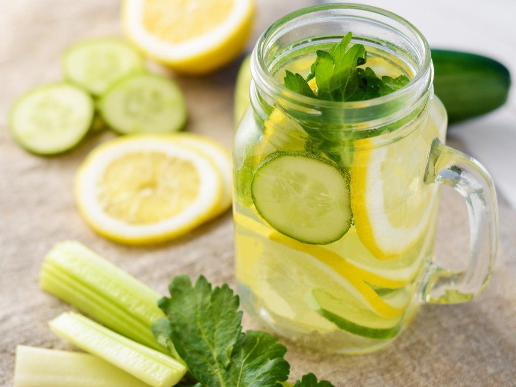 Glass mason jar of cucumber mint and lemon infused water on ice next to fresh summer celery — spa day refreshments at home]