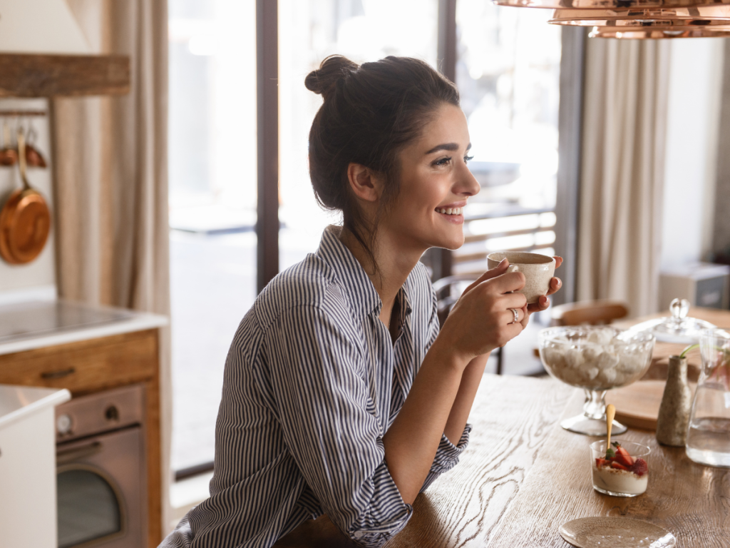 Woman sitting at a bright kitchen table with coffee and fresh fruit, slow summer morning routine at home]