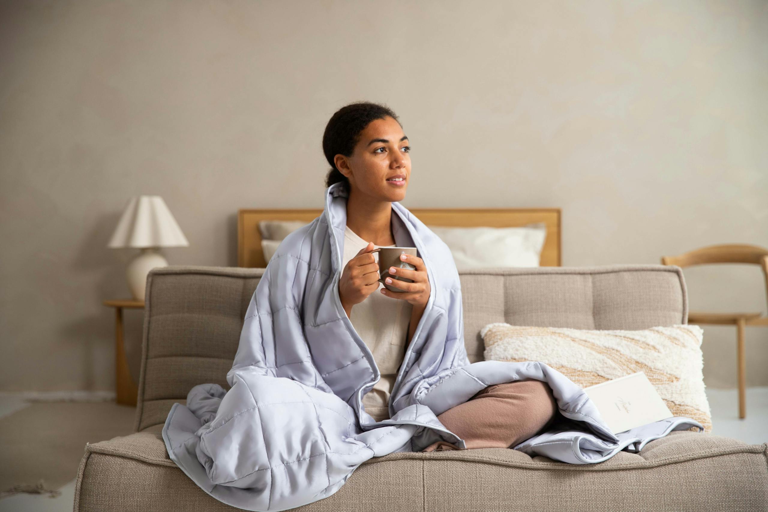 Woman in a Weighted blanket sitting cross-legged, enjoying coffee in a serene bedroom setting.