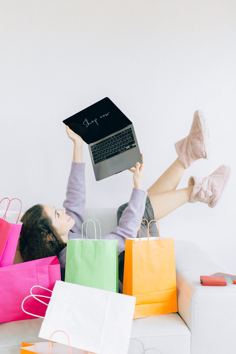 Woman lying on couch with laptop and colorful shopping bags, embodying online shopping joy.