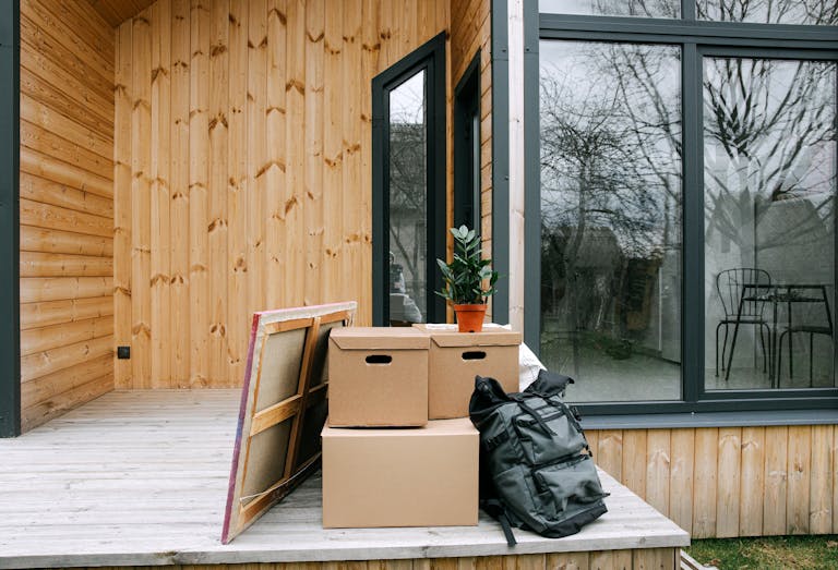 Wooden porch with cardboard boxes and potted plant next to glass windows. Moving into new home