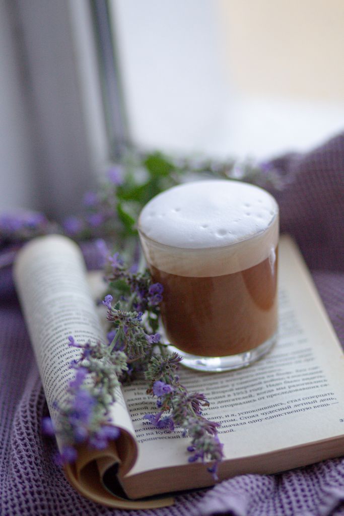 A warm cappuccino on an open book in a cosy reading nook surrounded by lavender flowers and a blanket.