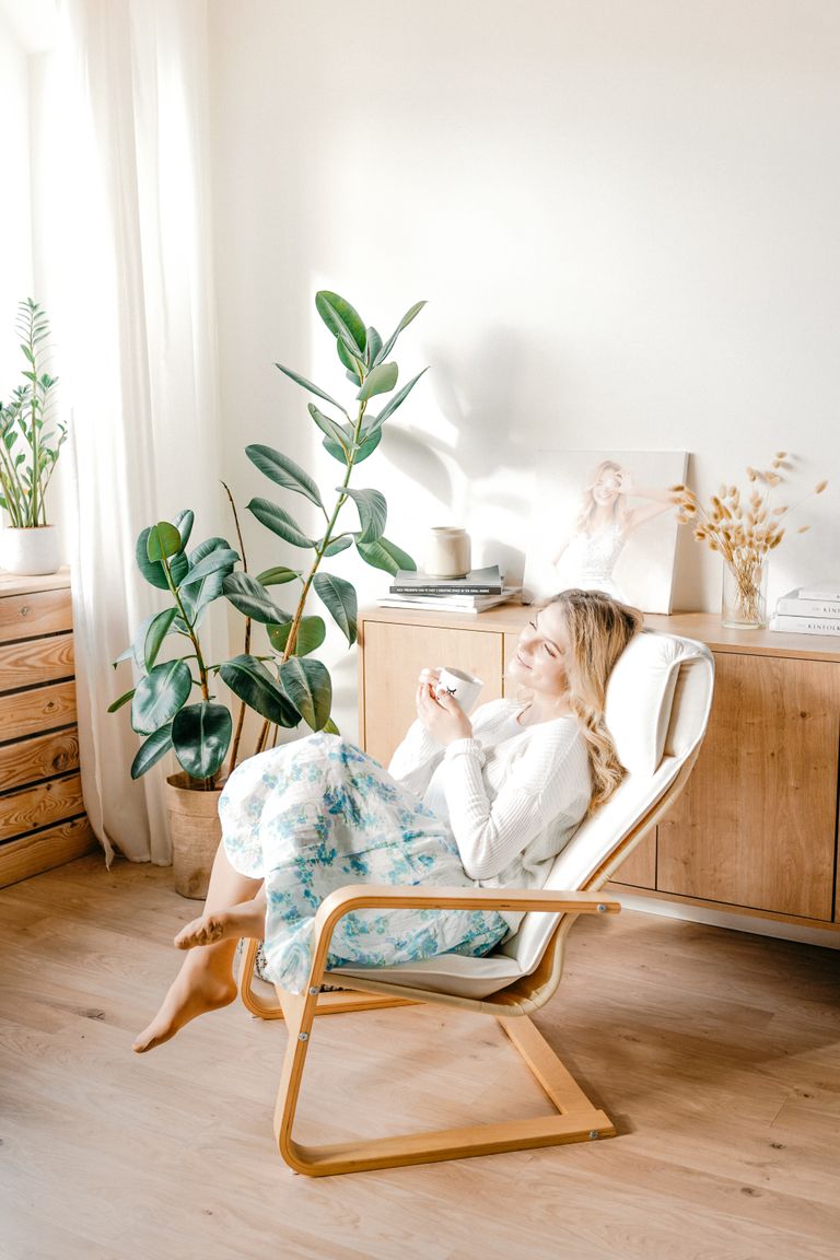 A woman enjoys a relaxing moment in a sunny, modern living room with plants and wooden furniture.
