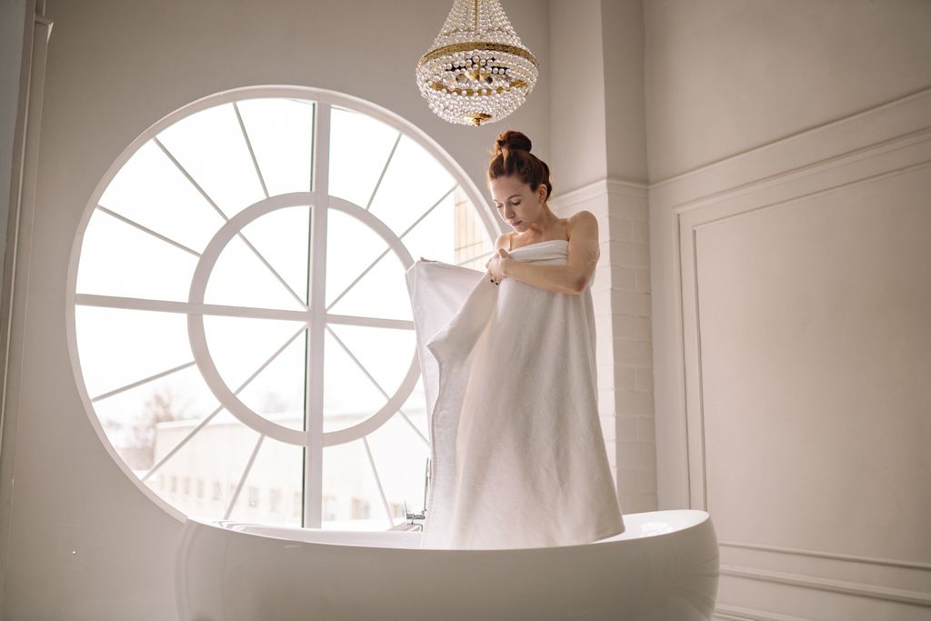 A woman wrapped in a towel stands elegantly in a bathtub by a large window.