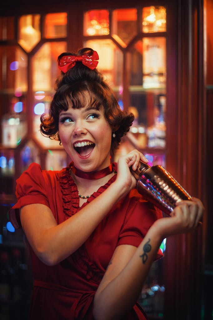 Cheerful woman in red dress joyfully shakes cocktail shaker in festive setting, capturing lively summer holiday spirit.
