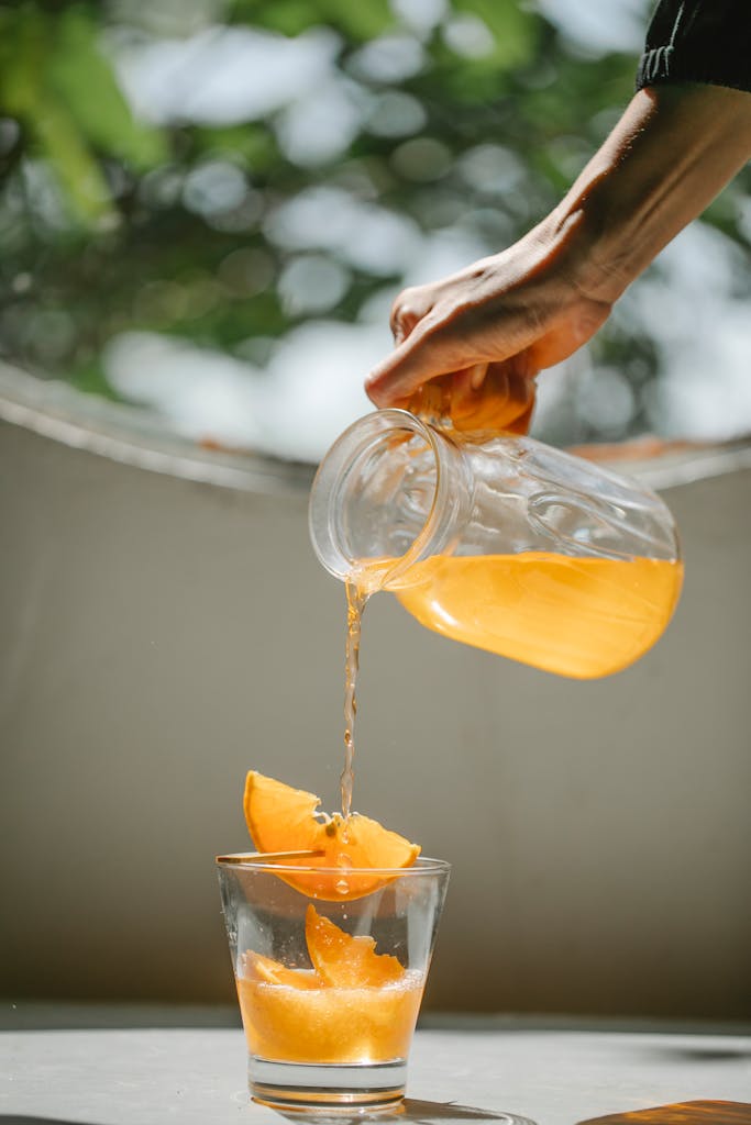 Crop anonymous woman holding transparent jar while pouring fresh cold lemonade made from fresh oranges into glass placed on surface in sunlight, British Cordial drink