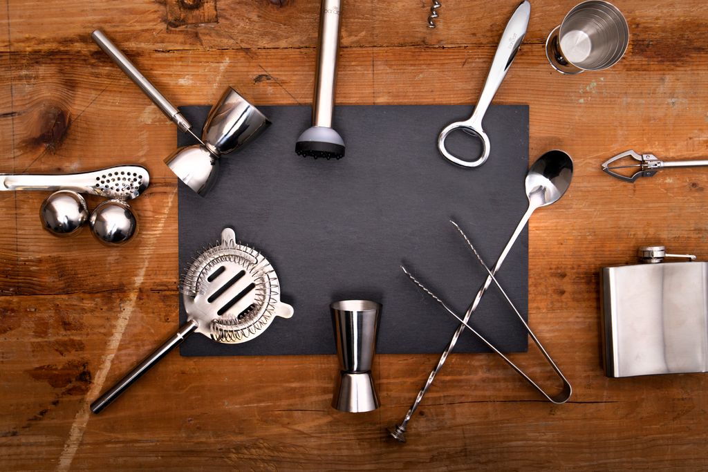 Flat lay of DIY home cocktail bar essentials including shaker, jigger, muddler and bar spoon on a wooden surface
