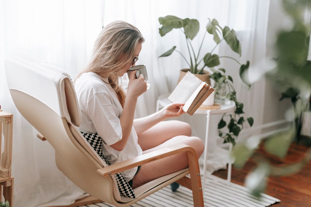 Side view of focused female drinking hot beverage while reading book in armchair in light room with potted green plants in a cosy summer Reading Nook