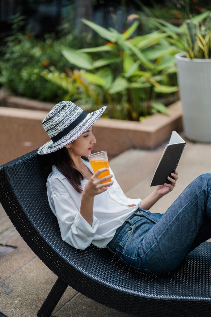 Side view of gorgeous young ethnic female tourist in stylish clothes and straw hat drinking natural juice and reading a novel while relaxing on sunbed in garden as part of a summer reading retreat at home