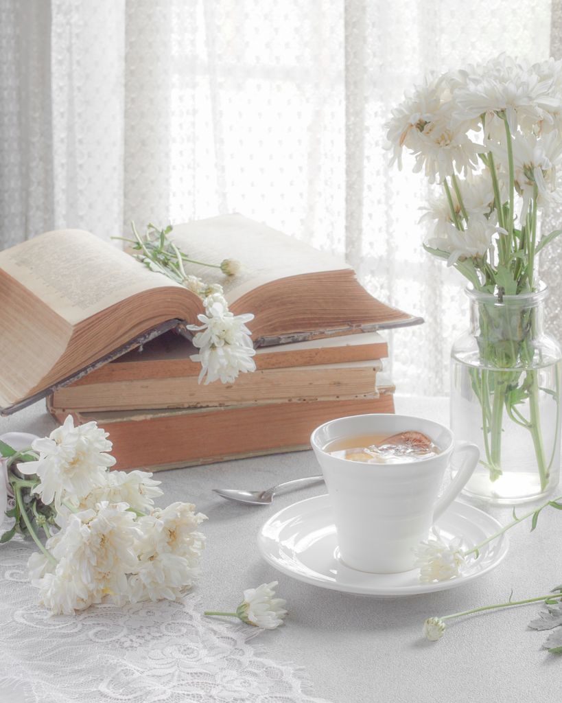 stack of classic summer books next to a glass of iced tea on a table for a reading retreat at home with White flowers on a lace tablecloth in soft morning light