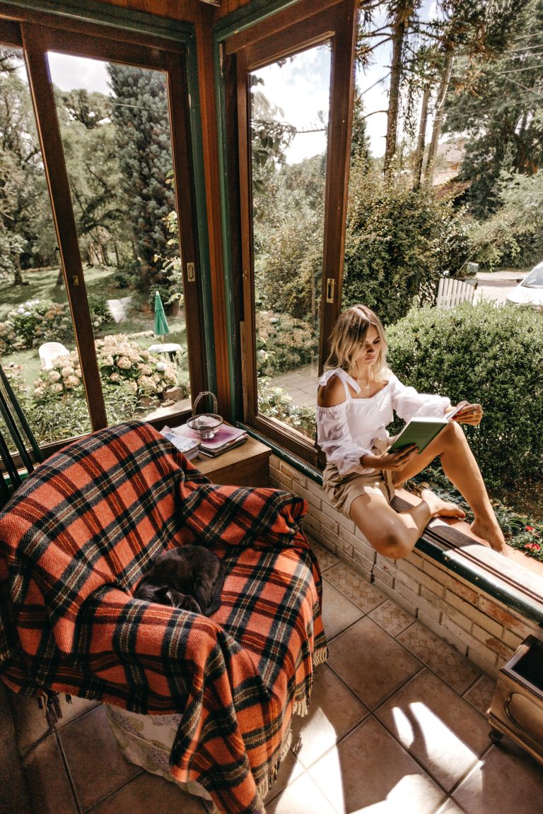 Woman enjoying a relaxing afternoon in a reading nook by a window with a garden view in summer