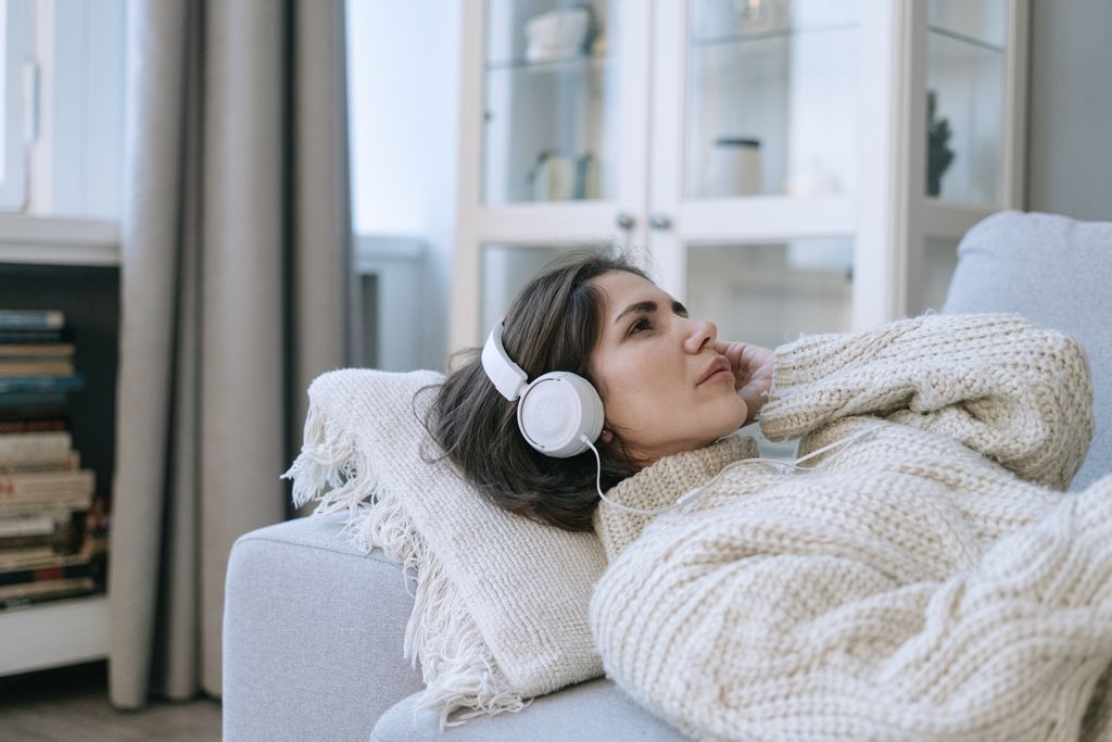 Woman in a knitted sweater relaxing on sofa, enjoying an audio book with headphones in an indoors reading nook