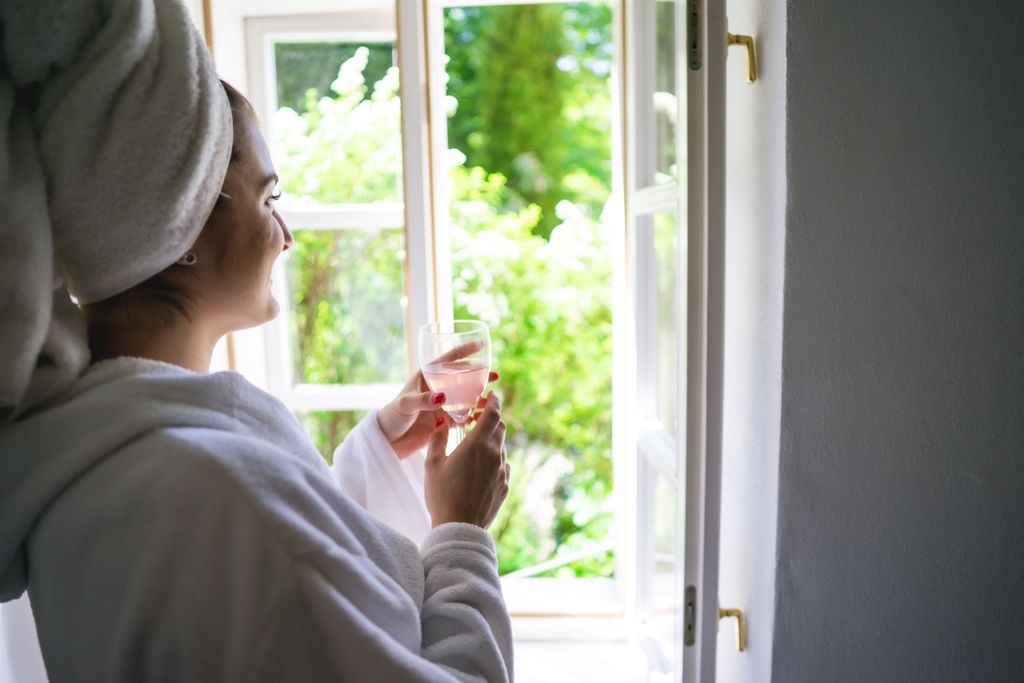 Woman in bathrobe enjoying a drink by the window, embracing a tranquil morning moment.