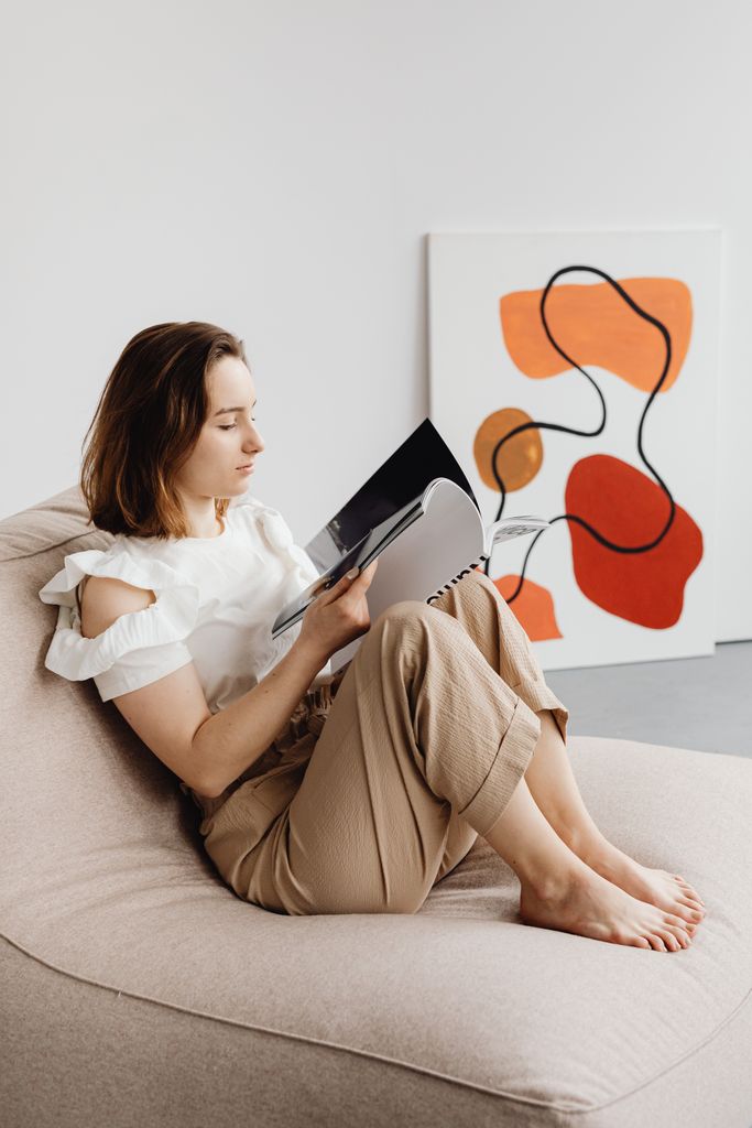 Woman reading a book in white blouse and beige pants reading indoors on a large cushion beside abstract art.