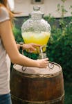 Woman pouring fresh lemonade from a dispenser on a wooden barrel in a sunny garden.