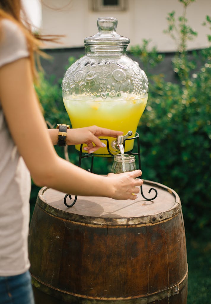 Woman pouring fresh lemonade from a dispenser on a wooden barrel in a sunny garden.
