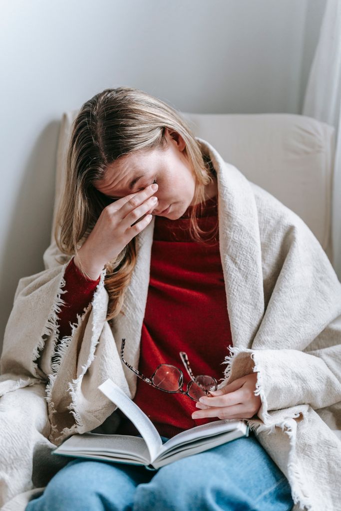 Woman sitting in an armchair with a blanket, holding a book and rubbing her eyes, falling asleep reading a book