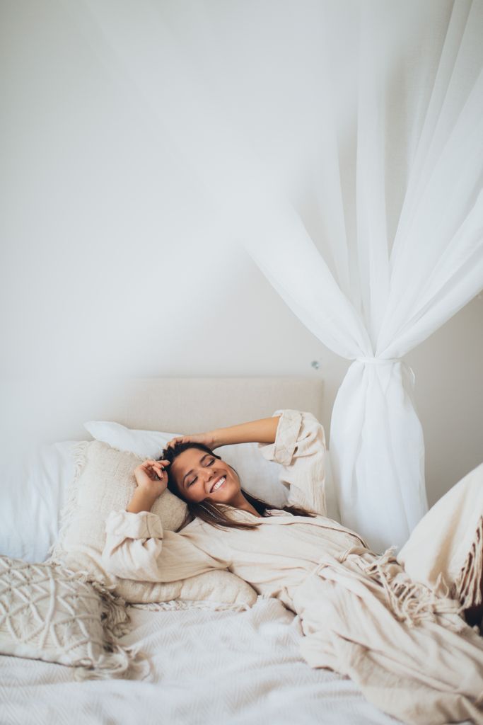 Woman smiling and relaxed in bed wearing a bathrobe in a cozy bedroom.