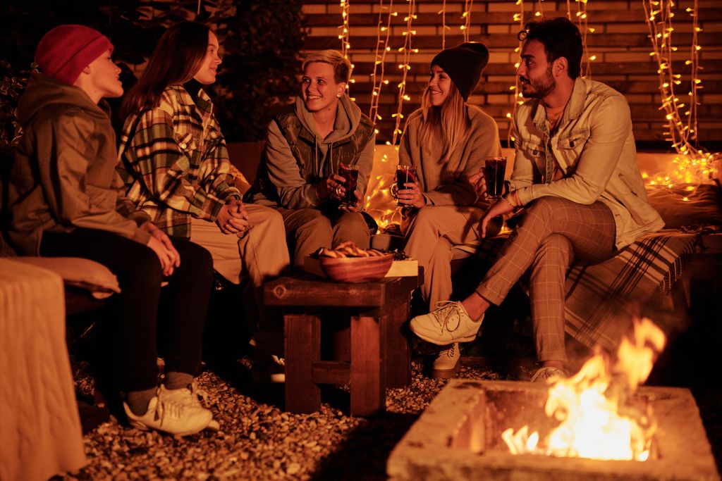 outdoor fire pit surrounded by a group of friends sitting on garden seating at night with fairy lights in background.