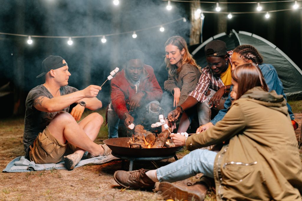 A group of friends relaxes by Marshmallows toasting over a small garden fire pit during a glamping staycation