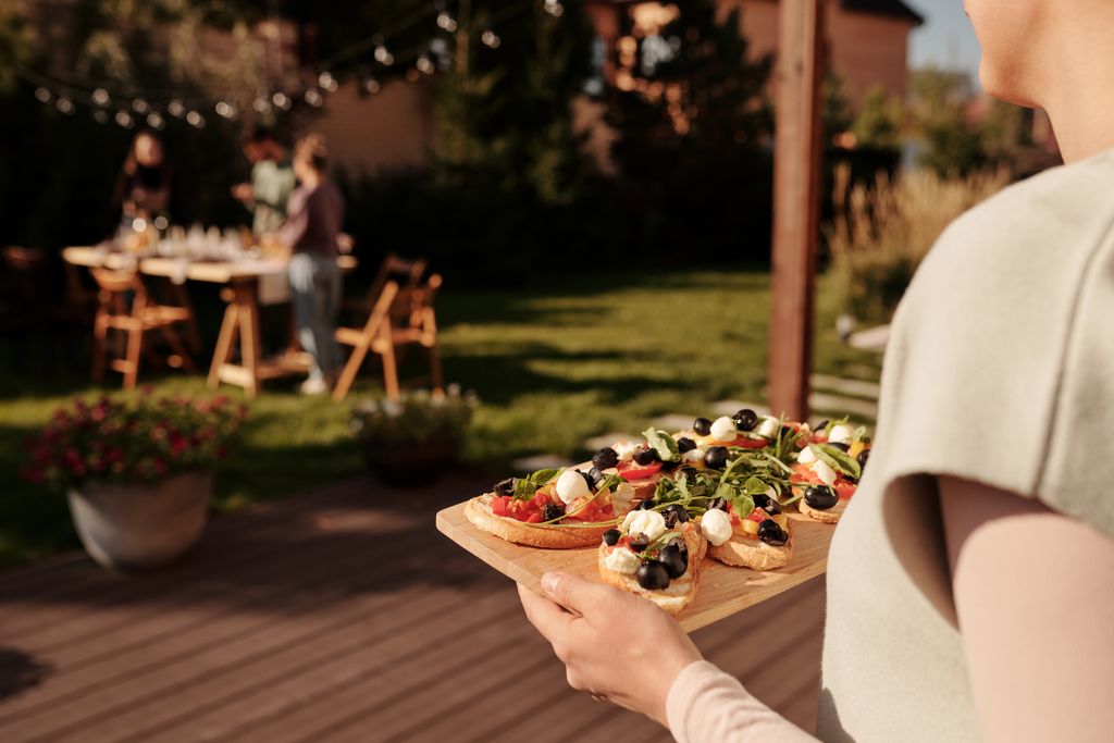 A woman holding a tray of fresh appetizers at an outdoor summer gathering with friends in a garden setting.