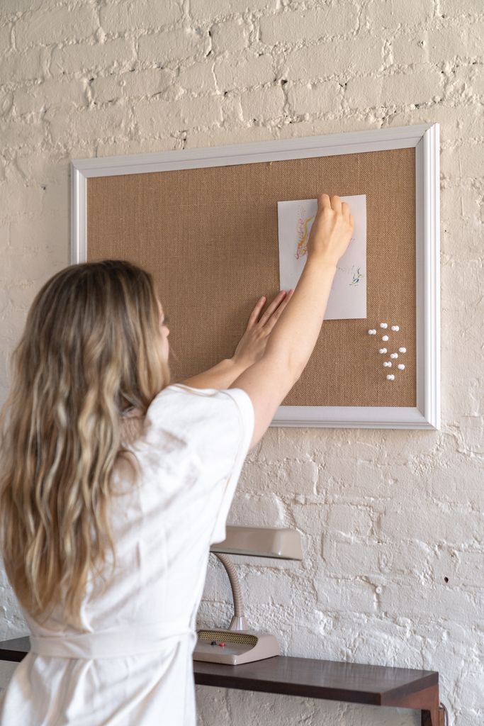 A woman places an artwork on a corkboard against a white brick wall.
