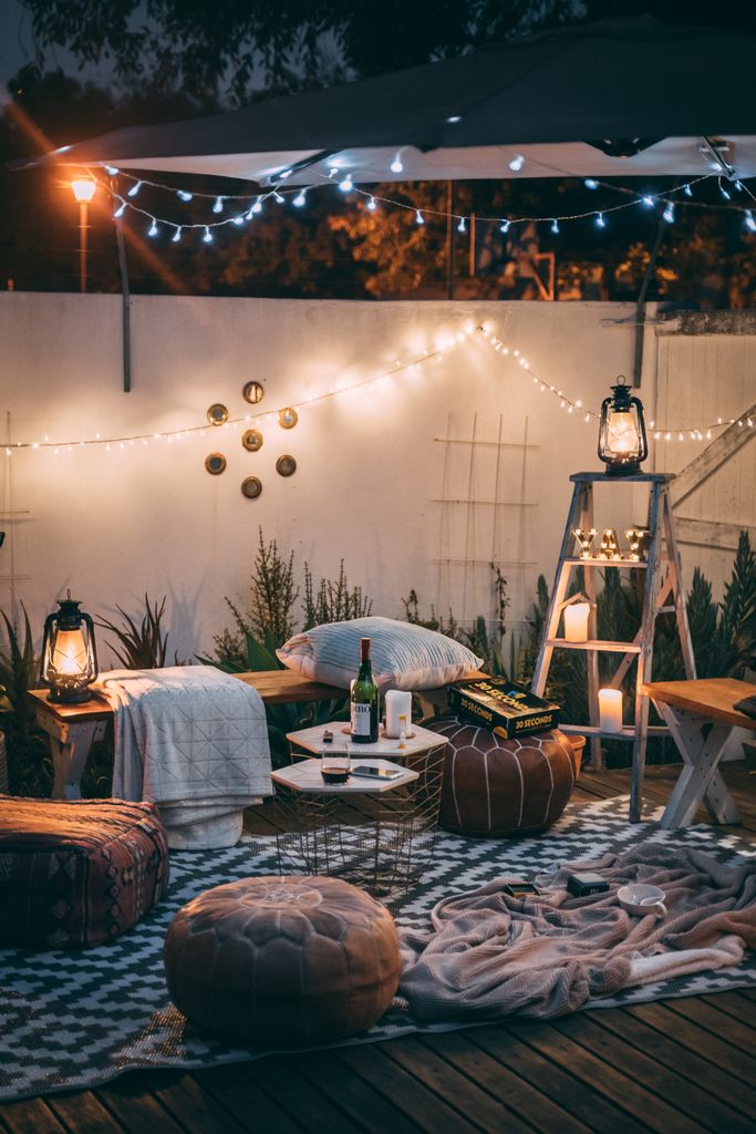 Charming outdoor patio setup for a glamping staycation with warm fairy lights and lanterns placed around benches and floor pillows.
