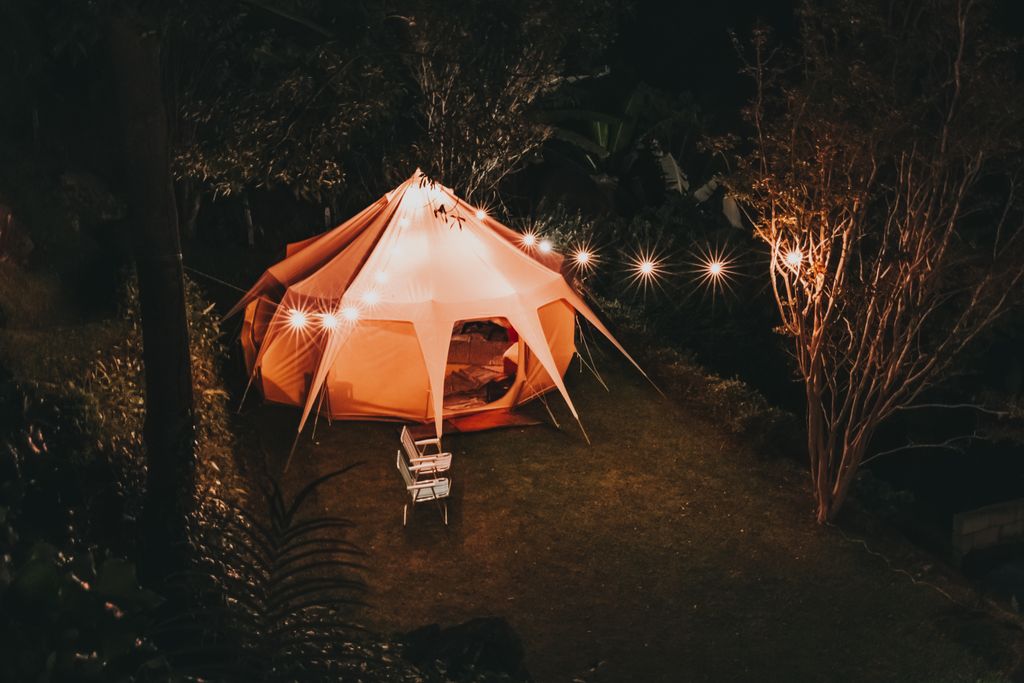 .Bell tent glowing at night in a UK back garden with fairy lights strung along a fence, garden glamping staycation setup